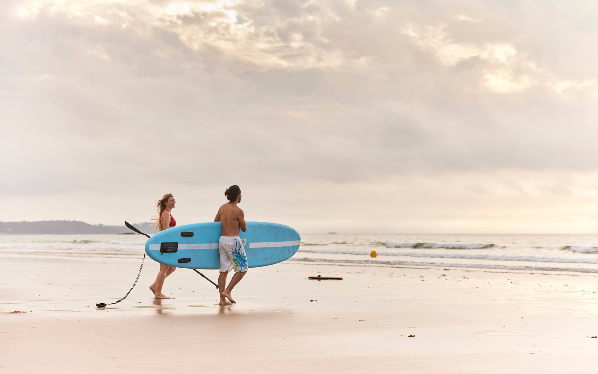 Deux adulte sur une plage marchant vers la mer, ils portent une planche de paddle